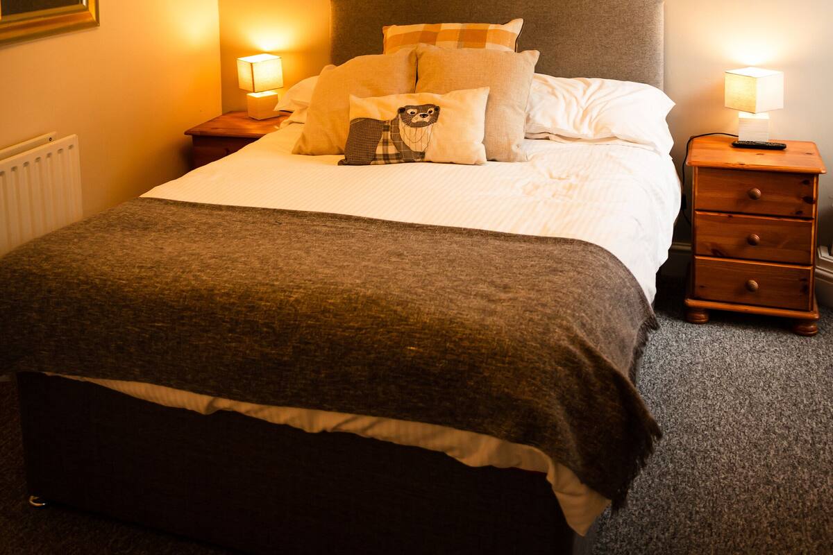 A neatly made bed with a headboard, two bedside tables with lamps, a hanging light fixture, and a radiator.