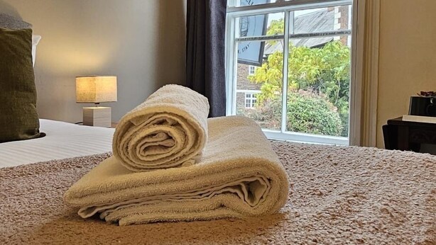 A neatly made bed with rolled towels, a window with a view of buildings and trees, and a lamp on a bedside table. - Cromwell's Tap House (Shrewsbury)