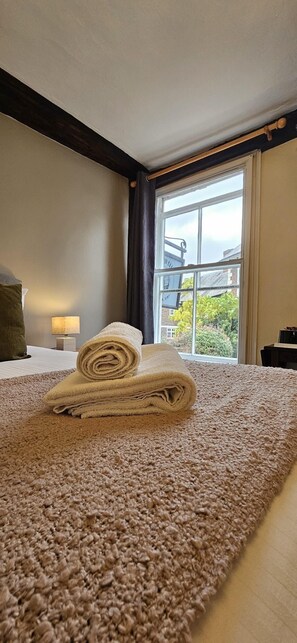 A neatly made bed with rolled towels, a window with a view of buildings and trees, and a lamp on a bedside table.