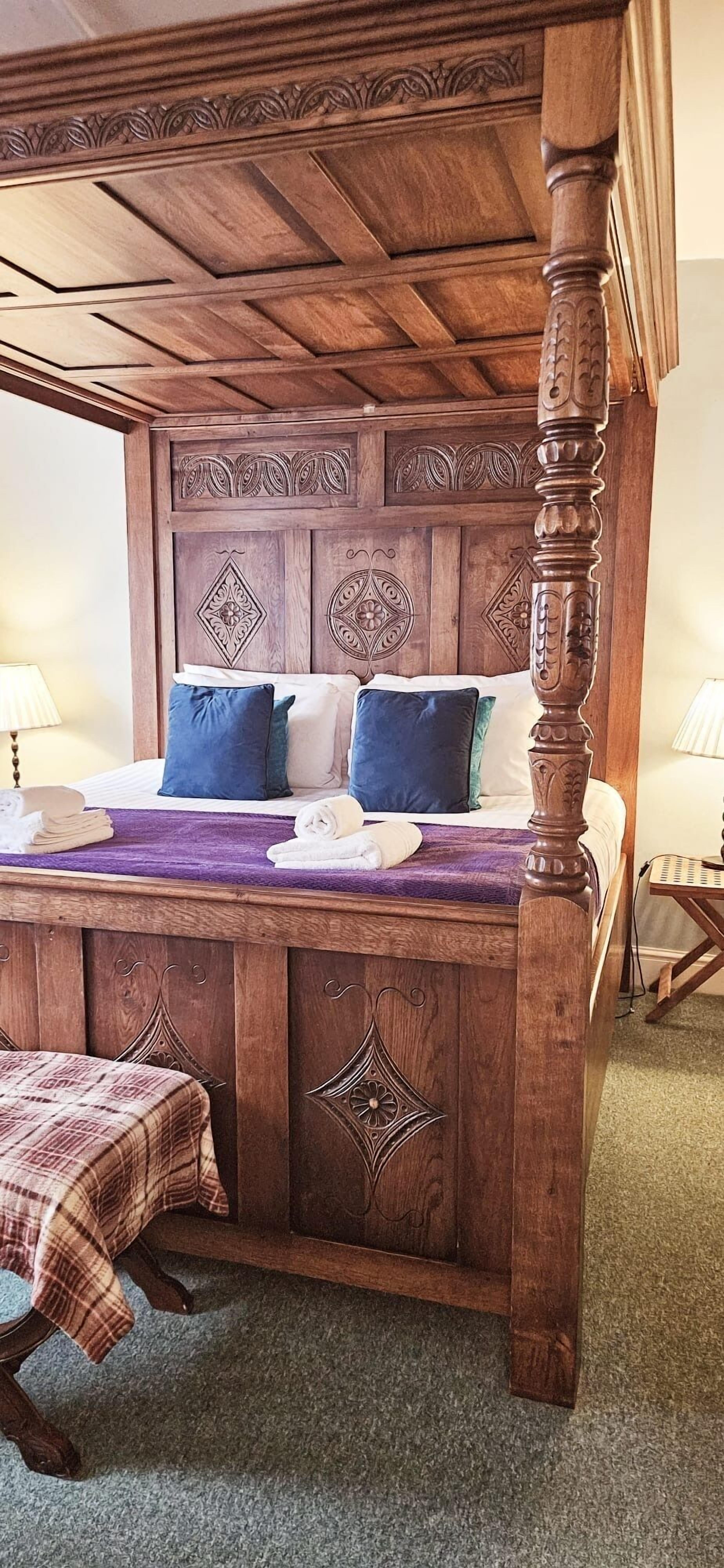 A four-poster bed with intricate woodwork, a purple throw blanket, and a plaid table runner.