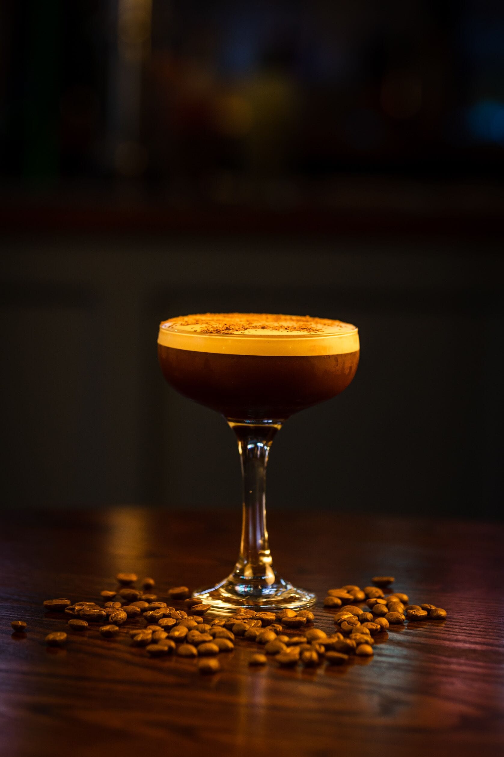 Un vaso de cóctel con una espuma en la parte superior, rodeado de granos de café dispersos sobre una superficie de madera.