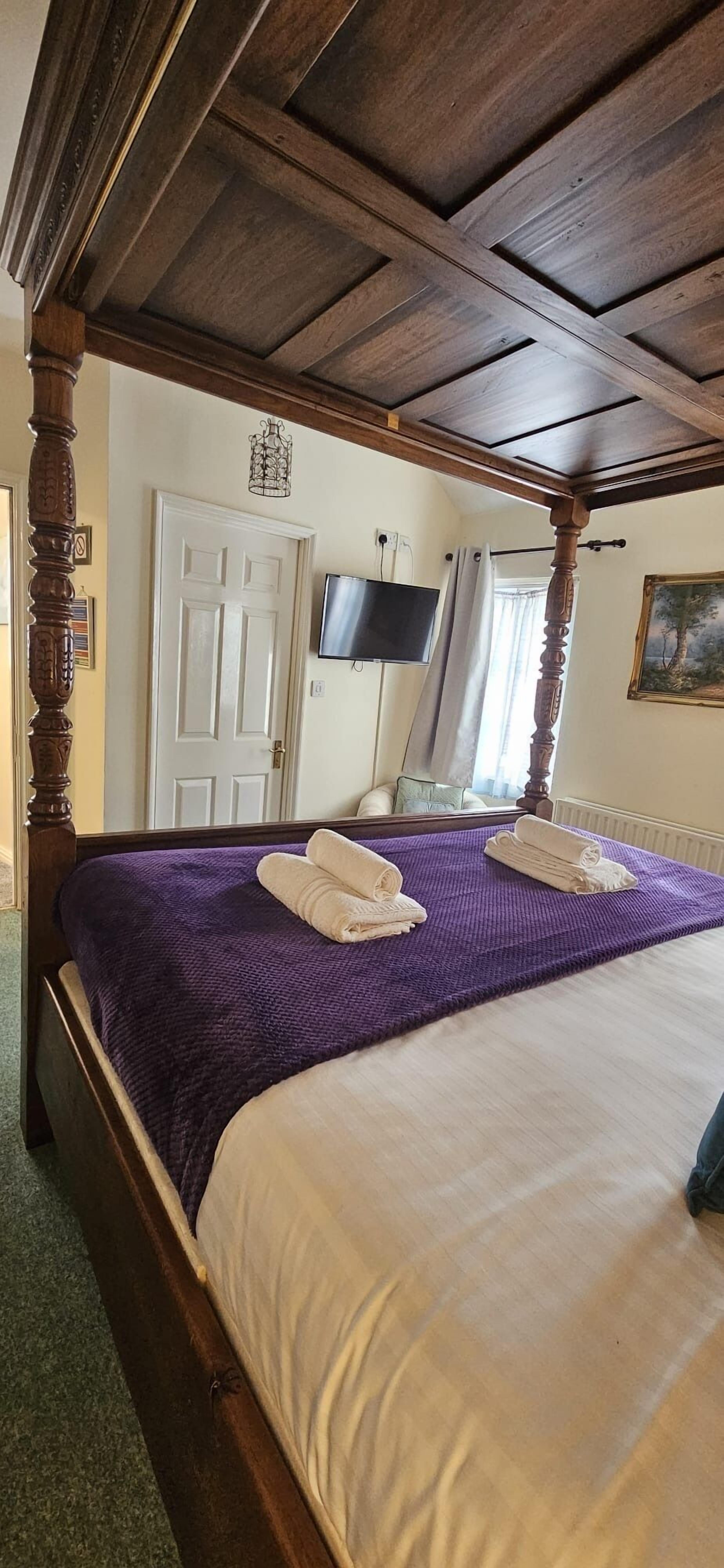 A four-poster bed with a purple blanket and white linens, a television on the wall, and a painting on the wall.