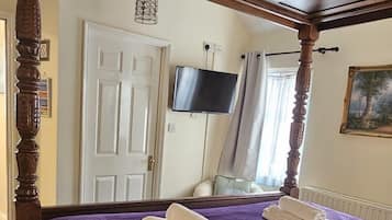 A four-poster bed with a purple blanket and white linens, a television on the wall, and a painting on the wall.