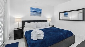 Una cama bien hecha con una colcha azul y toallas blancas, una lámpara de noche, una mesita de noche y un cuadro con una escena de playa en la pared.