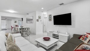Una sala de estar moderna con un sofá blanco, un sofá gris, una mesa de centro con un cuenco rojo, un televisor montado en la pared y una zona de comedor con una silla roja y una mesa blanca.