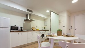 Een compacte keuken met een magnetron, fornuis en spoelbak. Er staat een eettafel met stoelen en een kleine potplant.