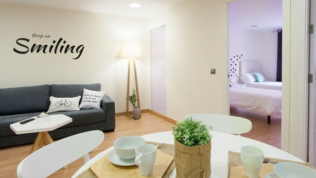 Een moderne kamer met een grijze bank, een eettafel met kommen en kopjes, een lamp en een bed op de achtergrond.