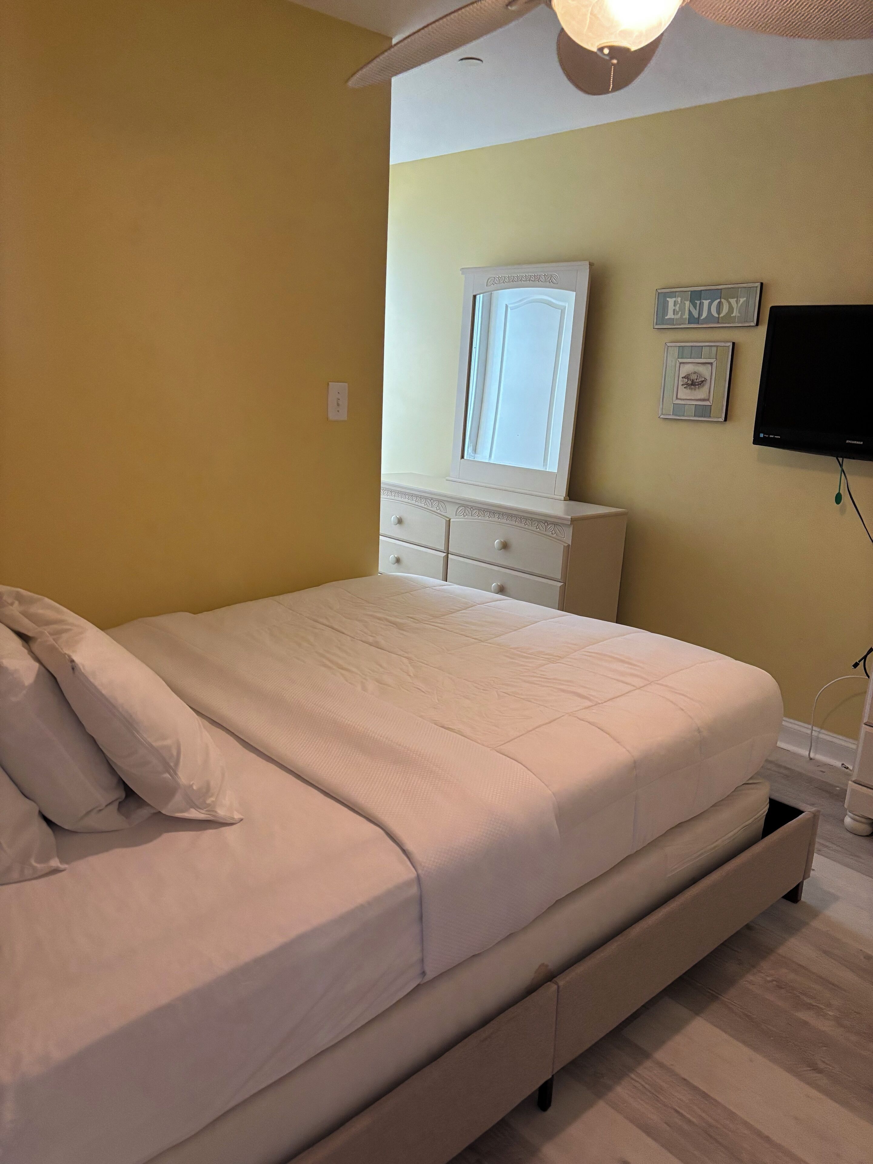 Un dormitorio con cama, cómoda, espejo, televisor y una pared con un letrero que dice 'ENJOY'.