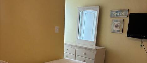 Un dormitorio con cama, cómoda, espejo, televisor y una pared con un letrero que dice 'ENJOY'.