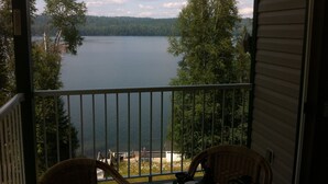 A balcony with wicker chairs, a small table, and a view of a lake surrounded by trees. - Birch Haven - A Luxury Lakefront Cottage On Pristine Horsefly Lake, with Wifi (Horsefly)