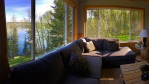 A cozy living room with a large window offering a view of a forest and lake. There is a sofa with patterned cushions, a small round table with a lamp, and a blue ottoman. - Birch Haven - A Luxury Lakefront Cottage On Pristine Horsefly Lake, with Wifi (Horsefly)
