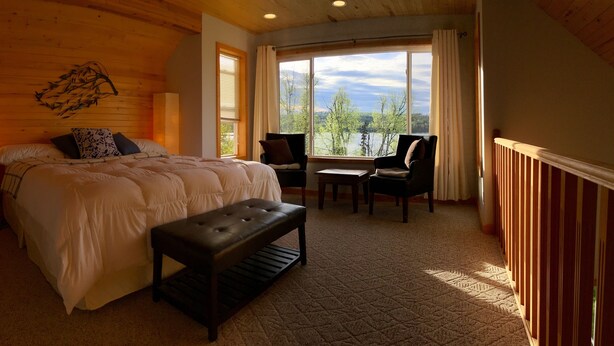A bedroom with a large bed, a bench, two chairs, and a view of trees and a lake through a window. - Birch Haven - A Luxury Lakefront Cottage On Pristine Horsefly Lake, with Wifi (Horsefly)