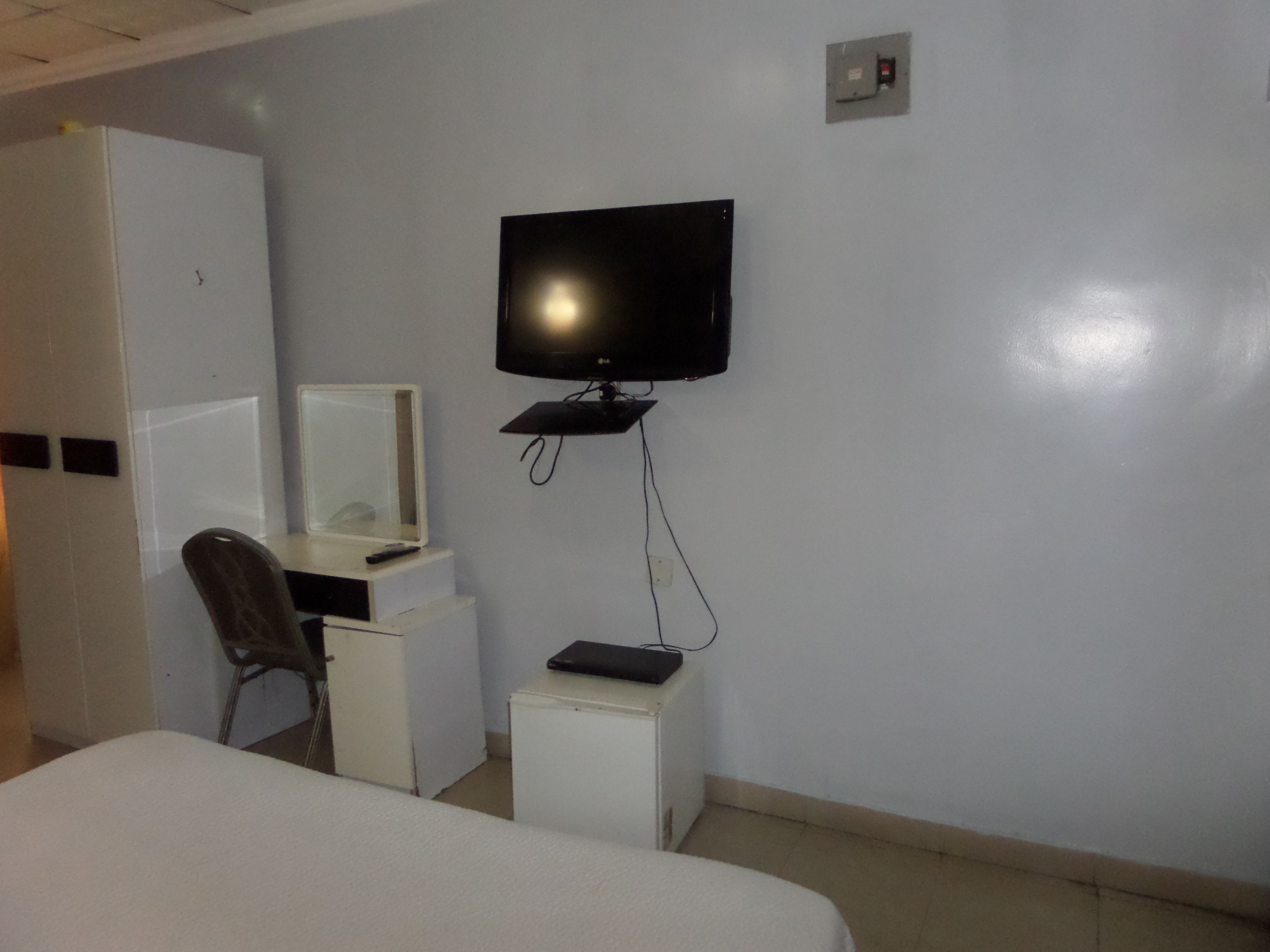A hotel room with a bed, a desk with a computer, a chair, a television mounted on the wall, and a small cabinet.