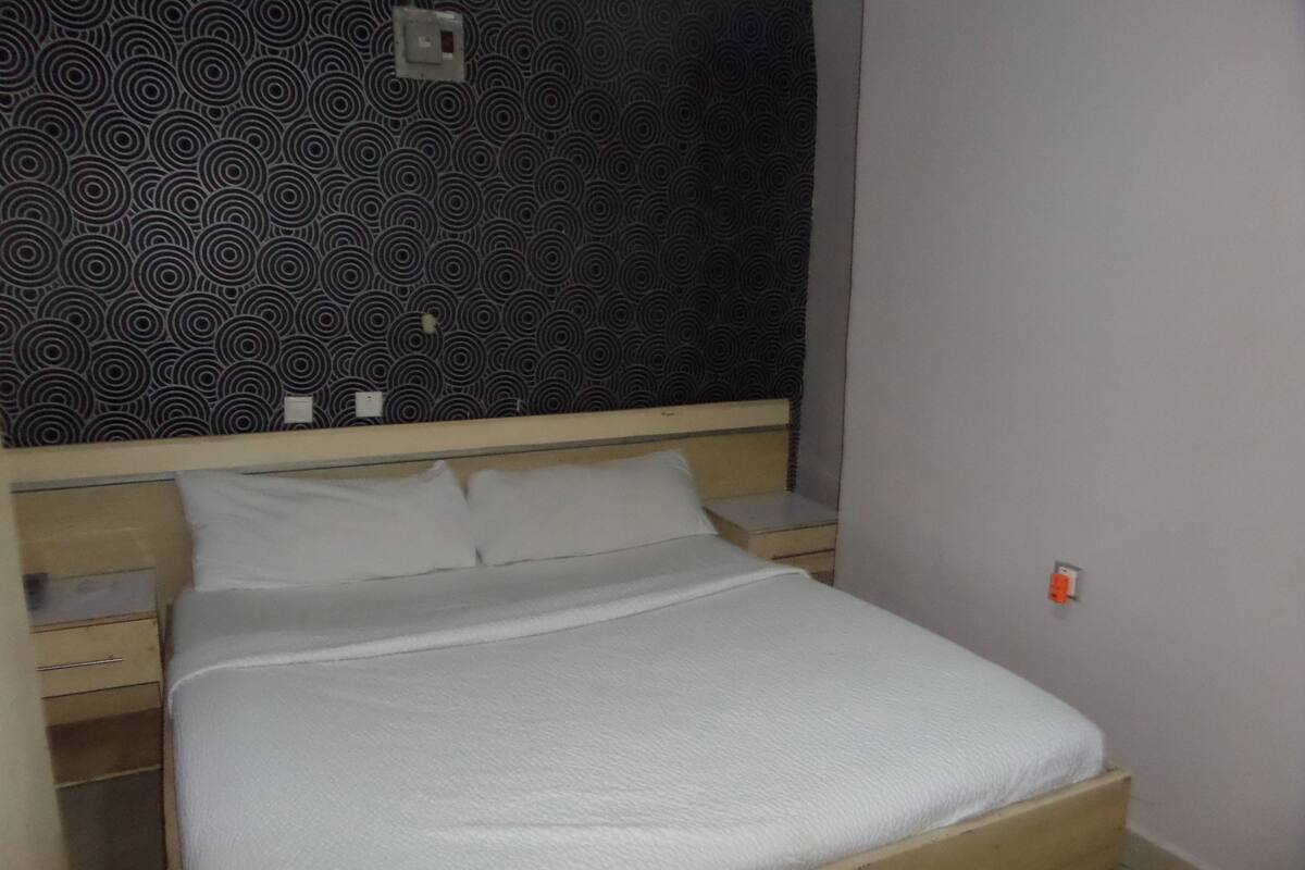 Una cama con estructura de madera y ropa de cama blanca, una mesita de noche y un papel tapiz estampado.