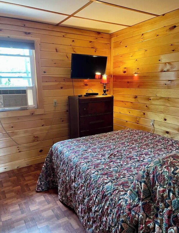 A wooden-paneled room with a bed, a dresser, a television mounted above it, and a window with blinds.