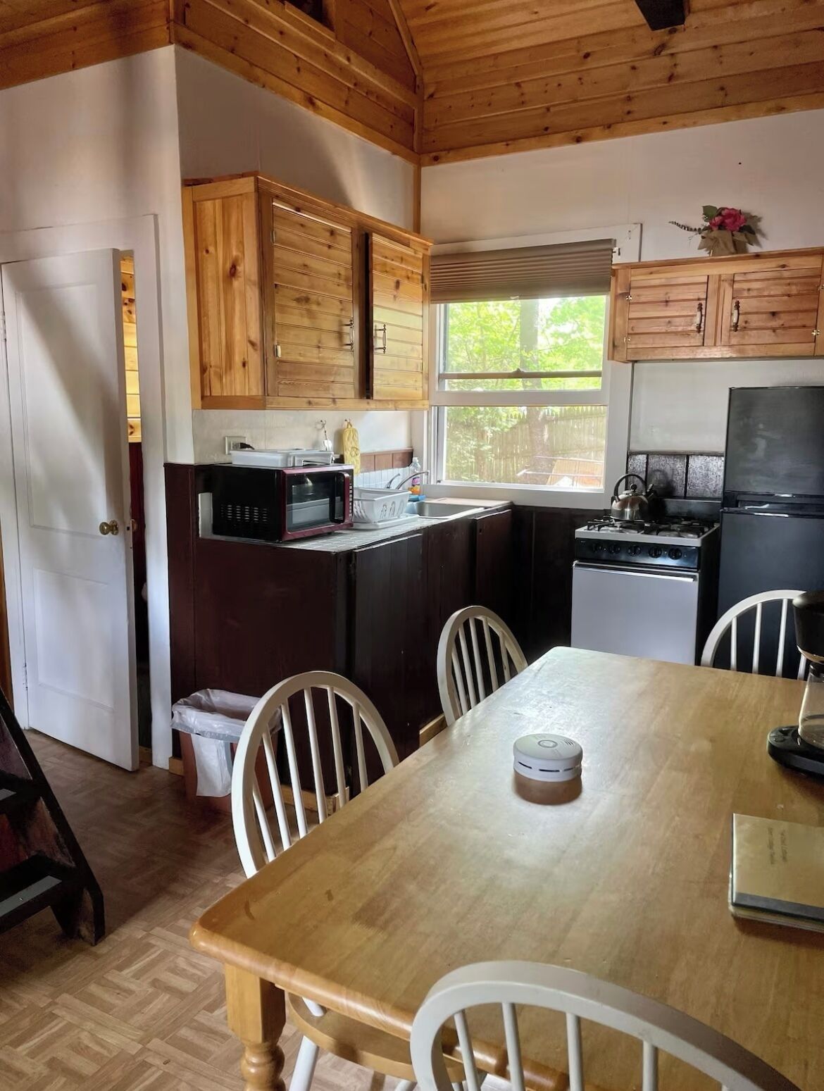 A compact kitchen with wooden cabinets, a microwave, a stove, and a sink. There is a dining table with chairs and a window providing natural light.