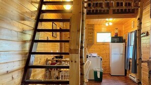 Interior de una cabaña de madera con escalera de madera, una zona de cocina con refrigerador y estufa, y una mesa de comedor con sillas.