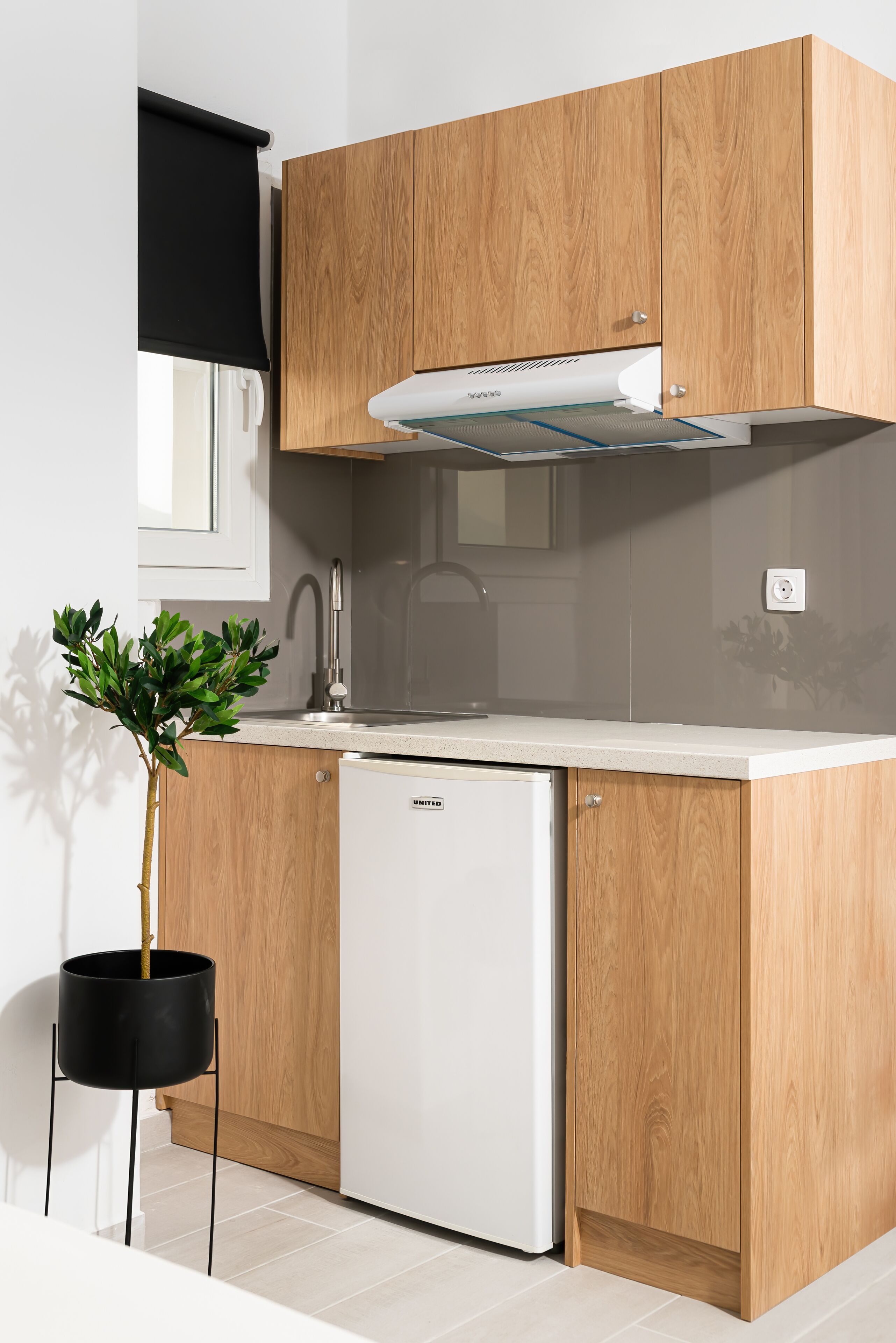 Une cuisine compacte avec des meubles en bois, un réfrigérateur blanc, une plante suspendue noire et une fenêtre avec un store noir.