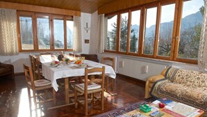 A cozy dining area with a wooden table set for four, surrounded by chairs. Large windows offer a view of mountains and trees. - B&B Marzola (Trento)