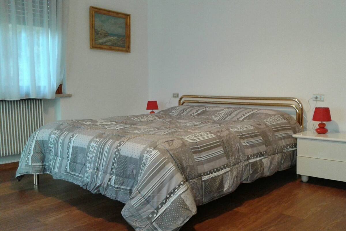 A bedroom with a bed, two bedside tables, a red lamp on each, a window with curtains, and a painting on the wall.