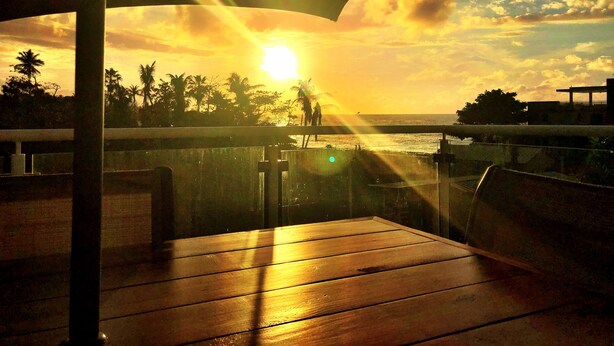 Une terrasse en bois offrant une vue sur le coucher de soleil sur l’océan, les palmiers et une côte lointaine.