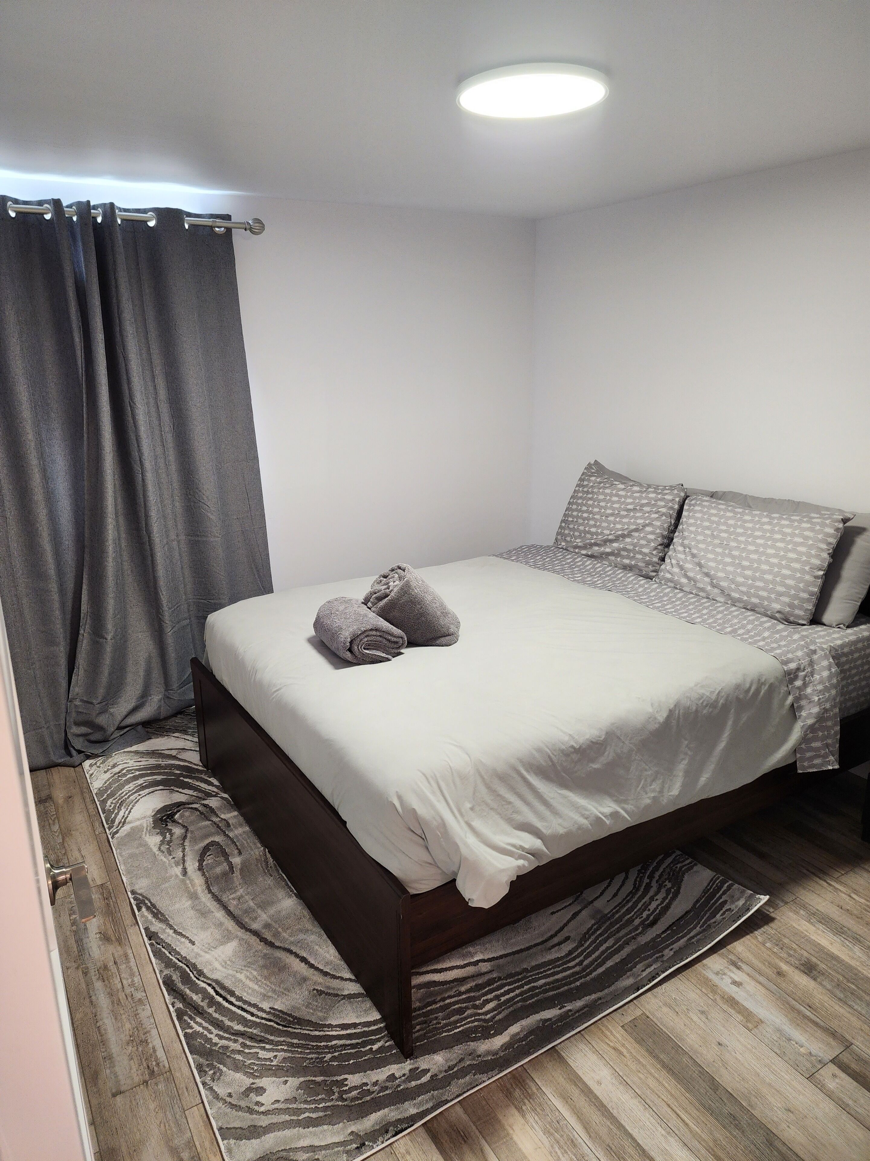A neatly made bed with a white bedspread and two pillows, a dark wooden headboard, a patterned rug, and a gray curtain.