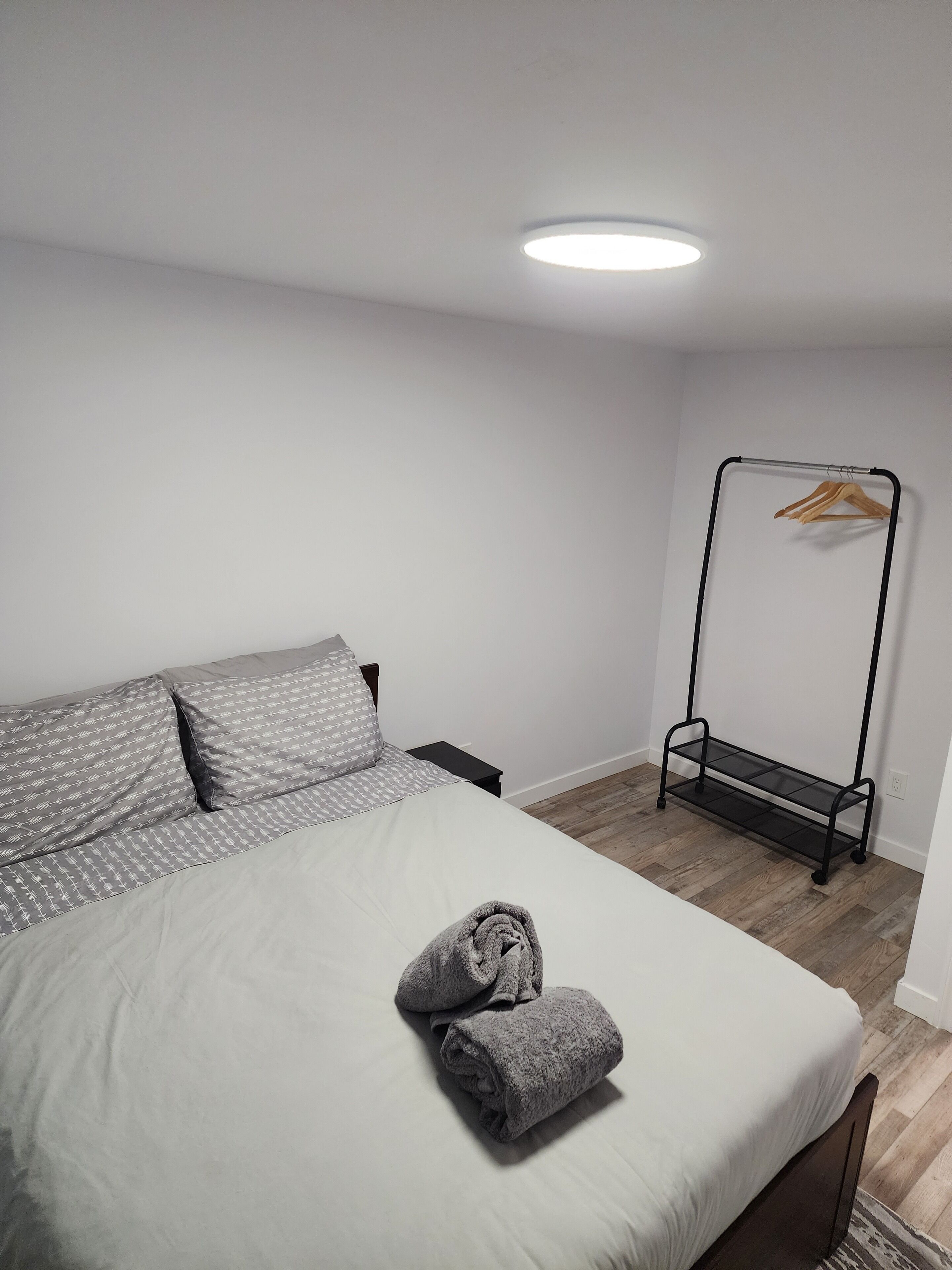 A neatly made bed with a grey blanket and pillows, a wooden hanger rack against the wall, and a small bedside table.