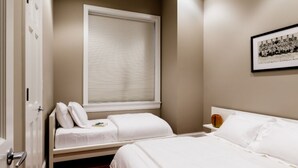 A bedroom with two beds, a window with blinds, a framed photo on the wall, and a bedside table with a cup.