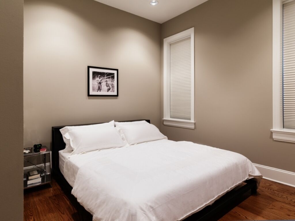 Een slaapkamer met een bed, een nachtkastje, een lijst met een foto aan de muur, en een raam met jaloezieën.