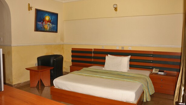A hotel room with a bed, bedside table, chair, and a painting on the wall. - Xcape Hotels and Suites Ltd (Calabar)