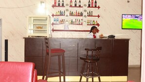 A bar area with a counter, shelves of bottles, two stools, and a television displaying a sports event.