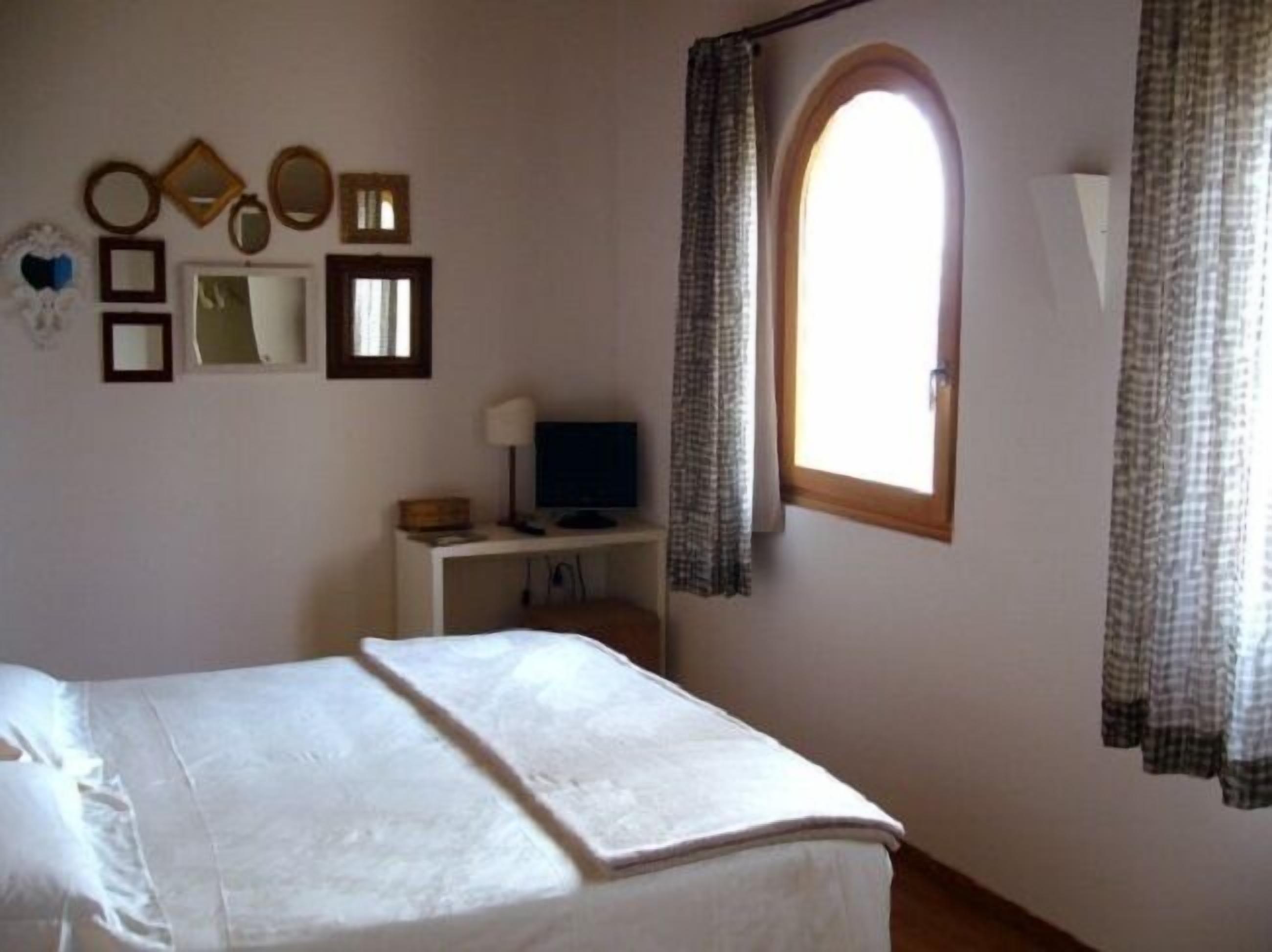 Ein Schlafzimmer mit einem Bett, einem Schreibtisch mit Computer, einem Fernseher und einem Fenster mit Vorhängen.