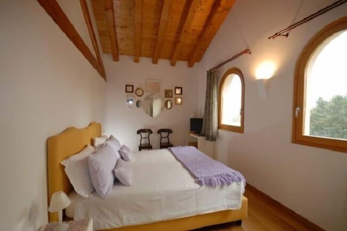 A bedroom with a wooden ceiling, a bed with white bedding and a purple throw, two bedside tables, a TV, and a window with a view of greenery.