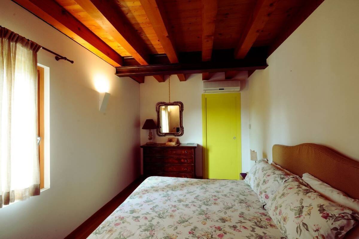 A bedroom with a wooden ceiling, a bed with floral bedding, a dresser, a mirror, and a yellow door.