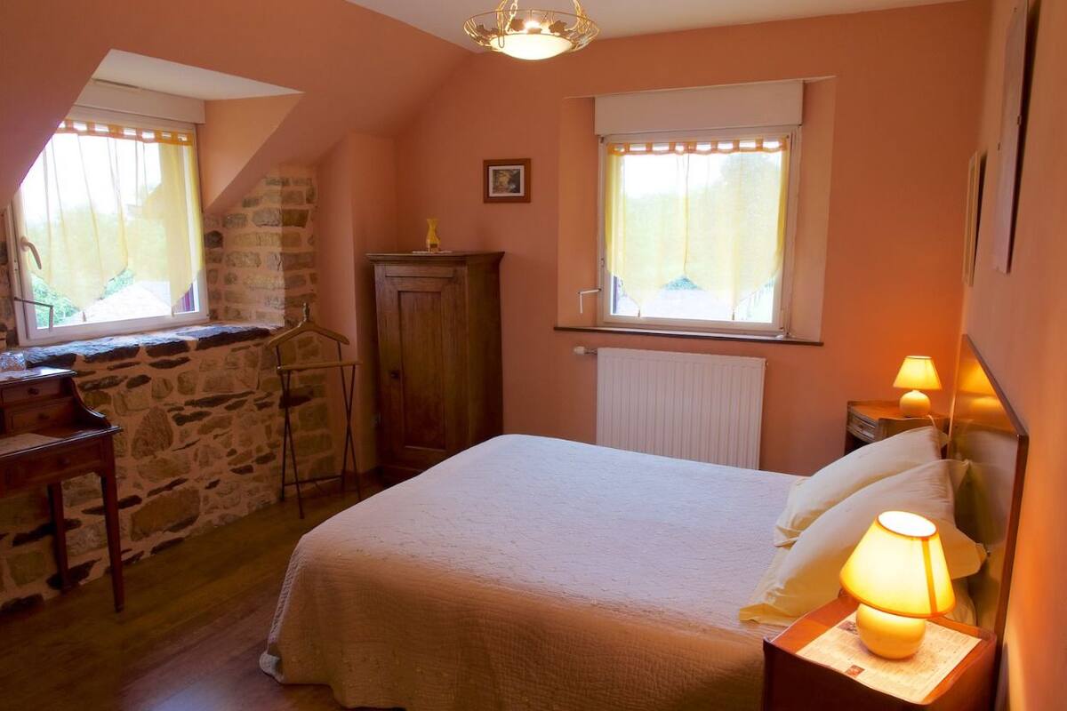 A bedroom with a bed, a nightstand with a lamp, a wooden dresser, a window with curtains, and a stone wall.