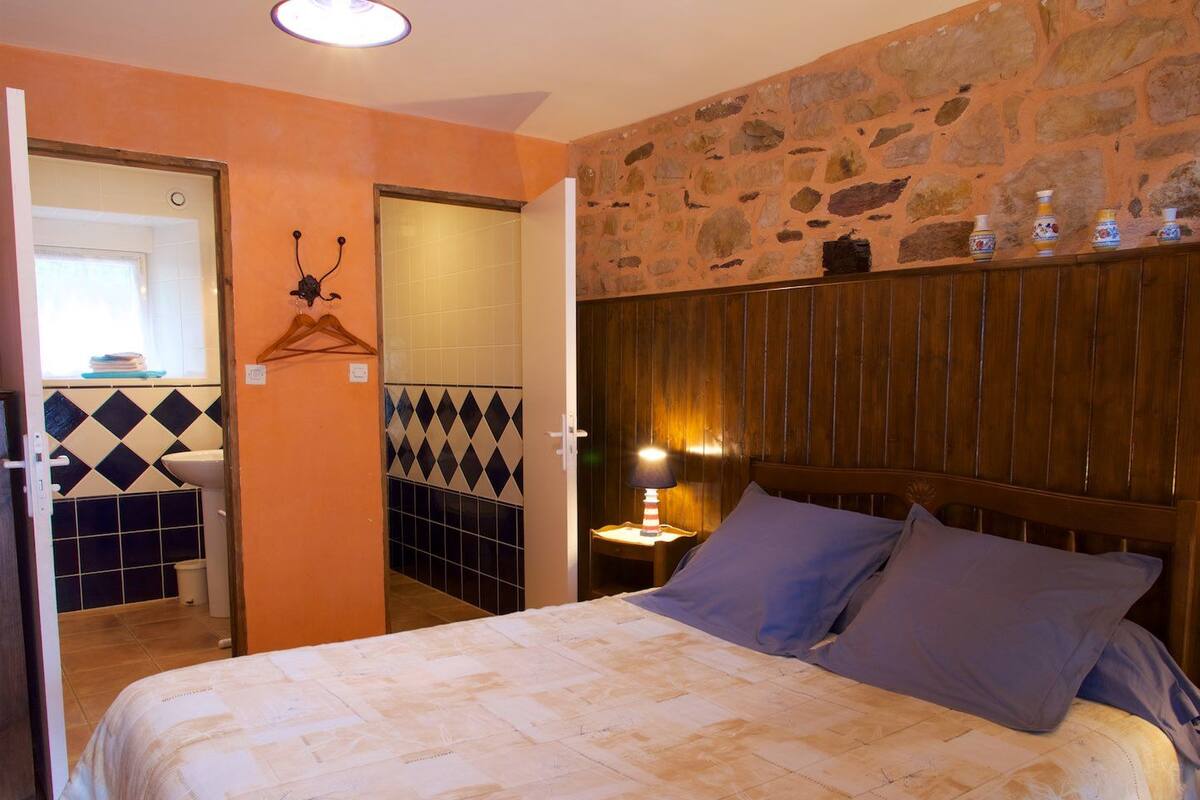 A bedroom with a bed, a nightstand with a lamp, a wooden headboard, and a stone wall.