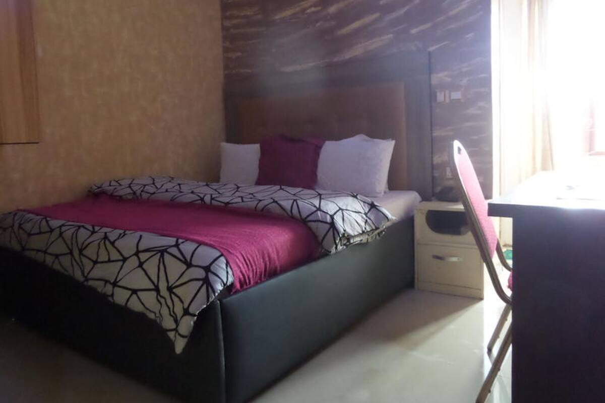 A neatly made bed with a patterned bedspread and pillows, a bedside table with a lamp, and a chair.