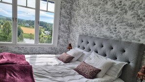 A bedroom with a large window, a bed with white linens and patterned pillows, and a grey upholstered headboard.
