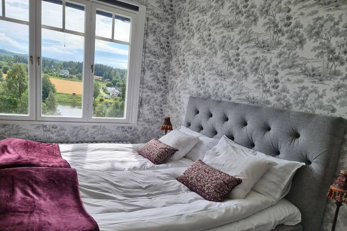 A bedroom with a large window, a bed with white linens and patterned pillows, and a grey upholstered headboard.