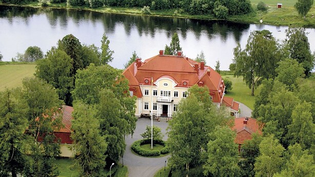 A large, two-story building with a red roof, surrounded by lush greenery and a lake.