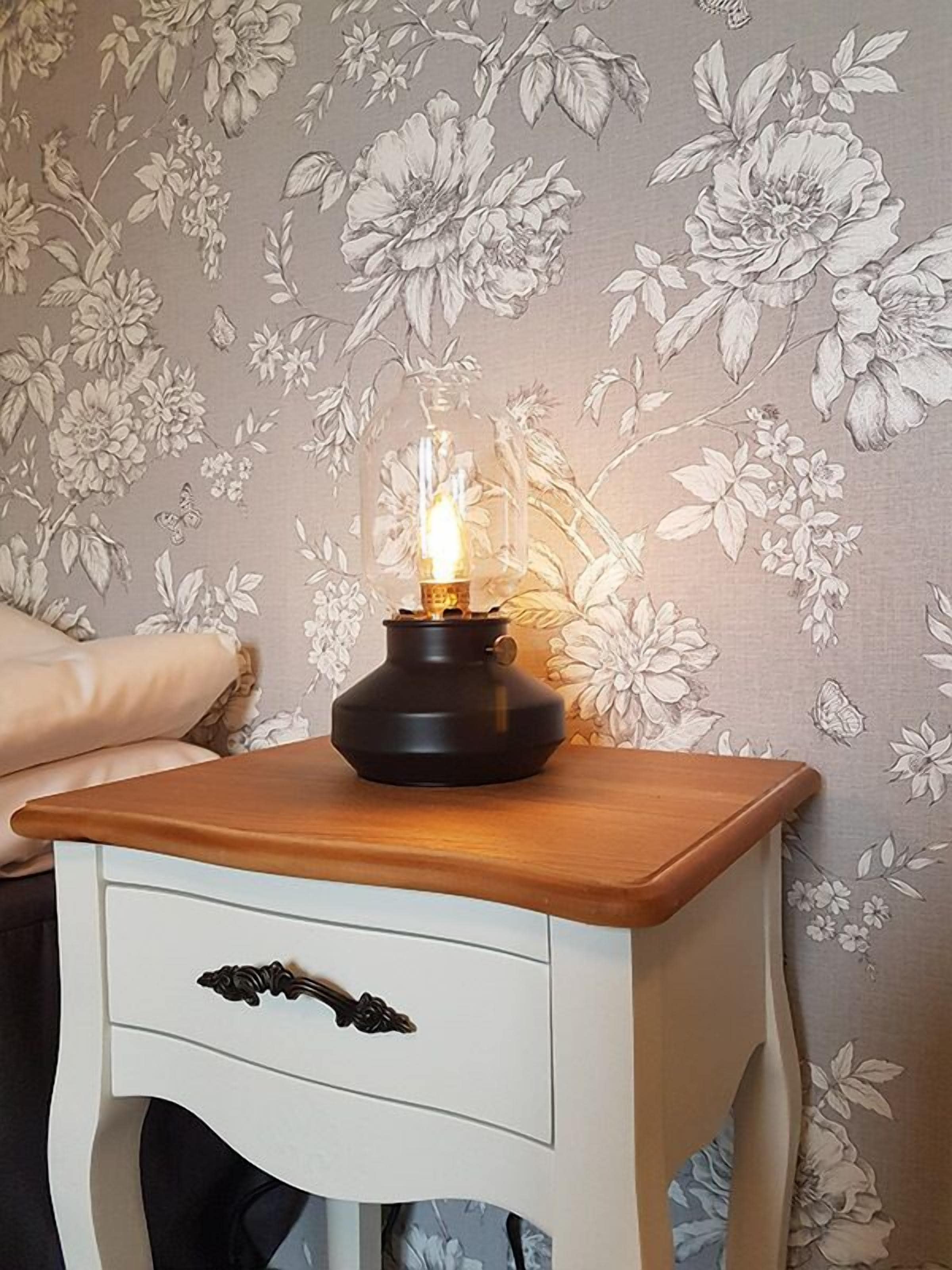 A bedside table with a lamp, a drawer, and a floral wallpaper background.