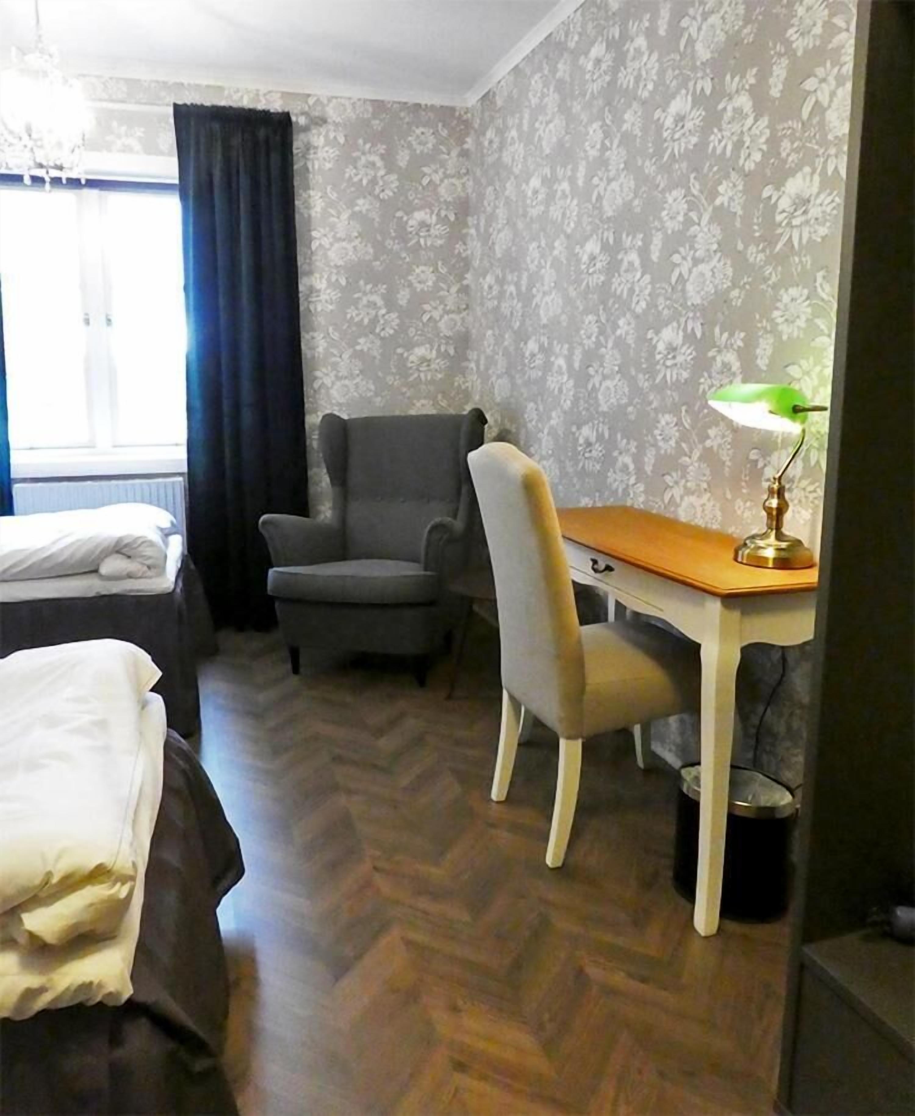 A hotel room with a bed, a desk with a lamp, a chair, a radiator, and a window with curtains.
