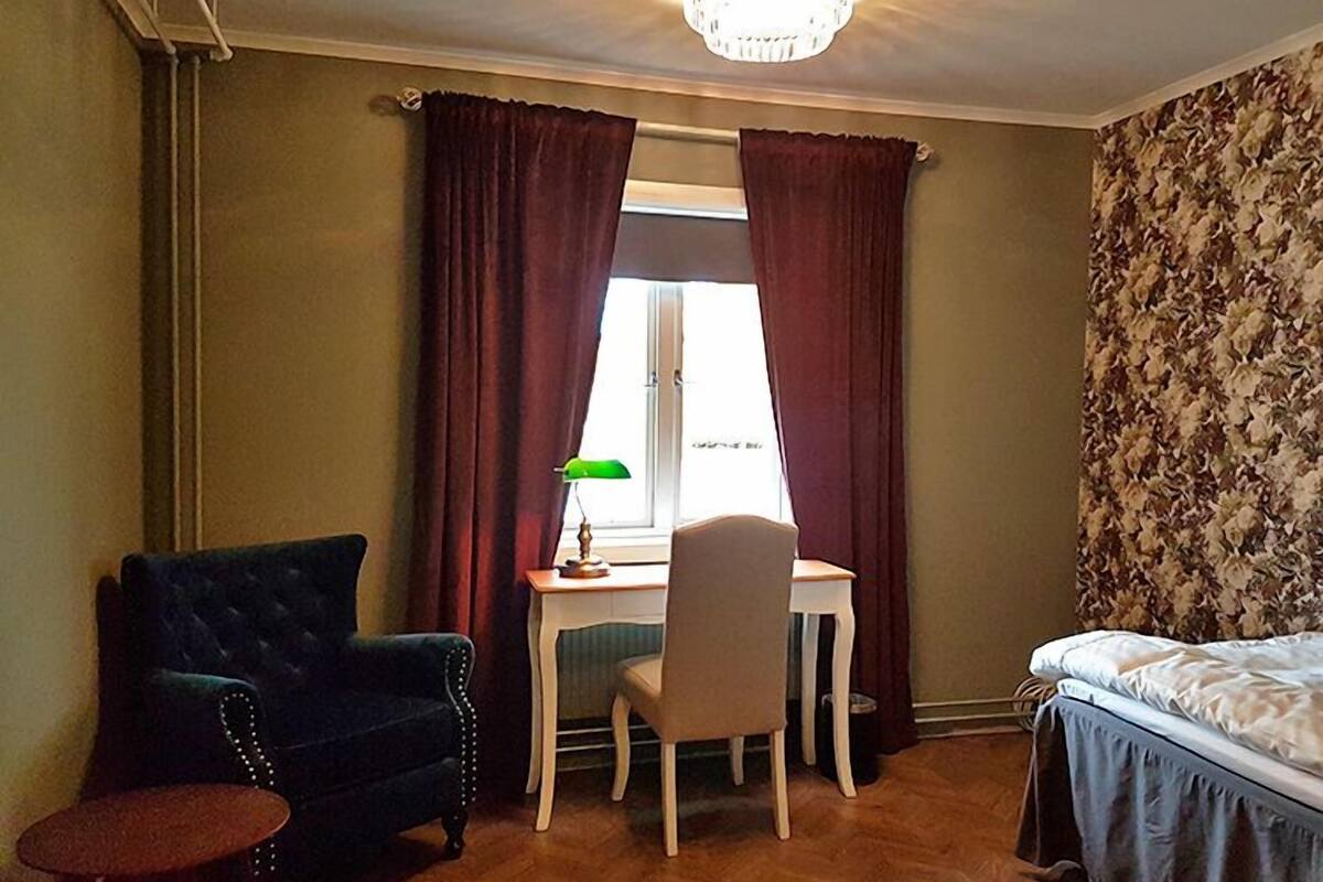A room with a desk, a chair, a small table, a bed, and a window with curtains.