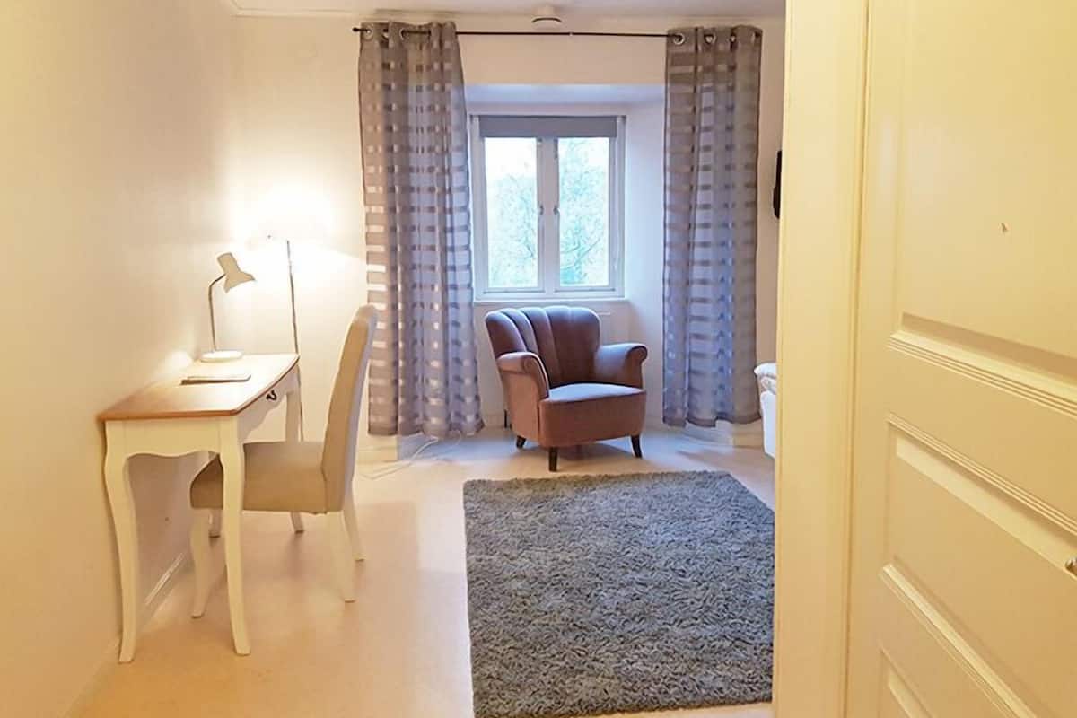 A room with a desk, chair, armchair, and a large window with curtains.