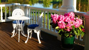 A wooden deck with white ornate chairs and a table, overlooking a lake and hills.
