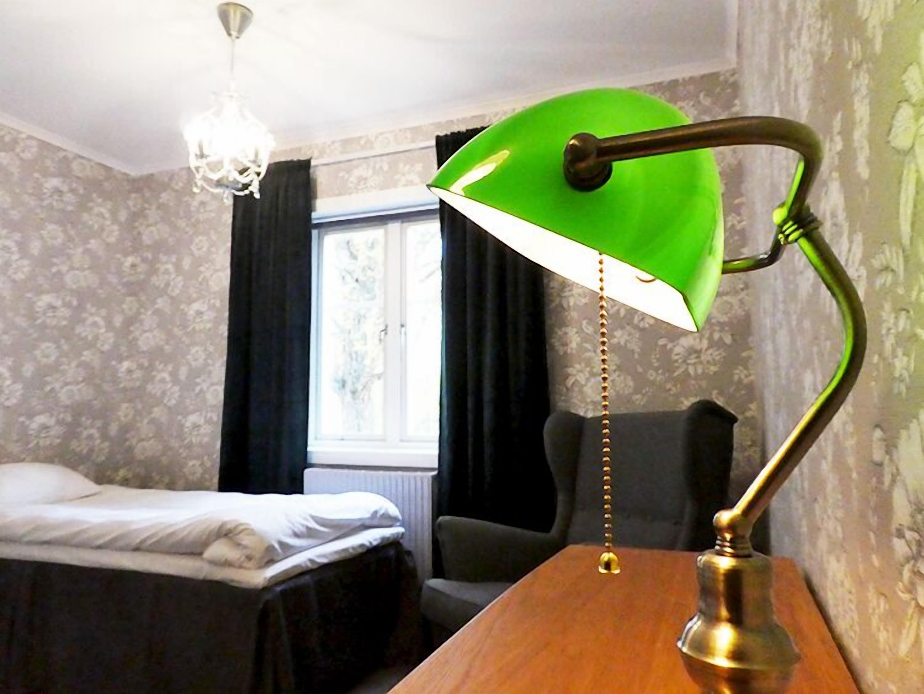 Ein Hotelzimmer mit Bett, Schreibtisch mit grüner Schreibtischlampe, Stuhl, Fenster mit Vorhängen und einem Kronleuchter.
