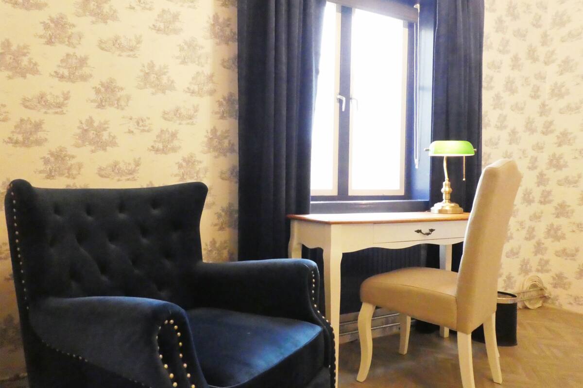 A room with a dark blue upholstered armchair, a white desk with a lamp, and a beige chair.
