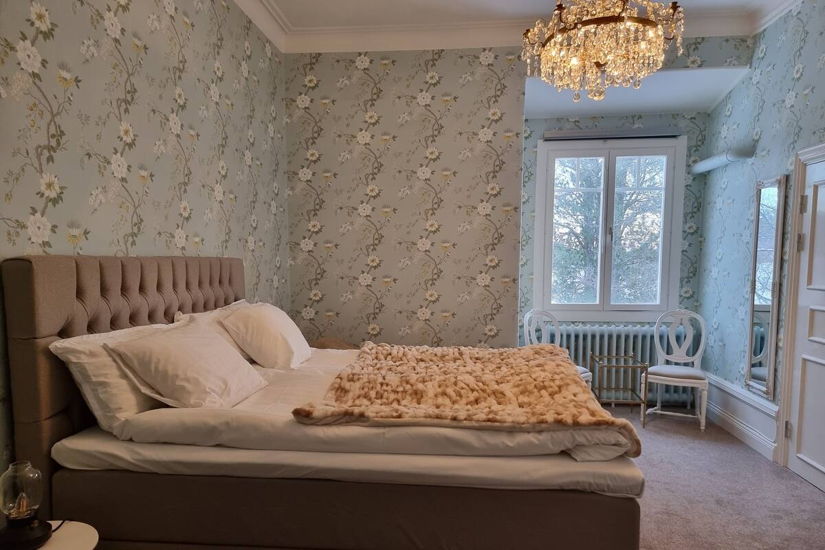 A bedroom with a tufted headboard, a large bed with white linens, a fluffy beige blanket, a white chair, and a window with a view of trees.