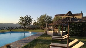 Un área junto a la piscina con sillones reclinables, una estructura de techo de paja y un edificio de piedra al fondo.
