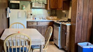 Una cocina compacta con armarios de madera, un microondas, una estufa y un lavabo. Hay una mesa de comedor con sillas y un cubo de basura azul.
